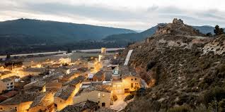 Fontanero Jorba trabajando cerca de una avenida arbolada con casas de piedra y paisaje montañoso de fondo.
